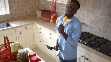 Mutfakta dans eden kameraya bakarak yemek pişirme aletinde şarkı söyleyen neşeli bir gencin yüksek açılı portresi. Mutlu Afrikalı Amerikalı adam evde yemek pişirip poz verirken eğleniyor..