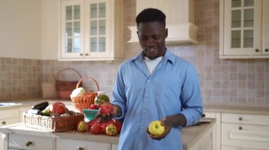 Afro-Amerikan pozitif adam organik vitamin elmalarıyla hokkabazlık yapıyor kameraya gülümsüyor. Mutfakta hasatla poz veren mutlu genç çiftçinin portresi..
