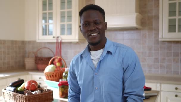 満足したアフリカ系アメリカ人の若者が、カメラを見て笑顔で保存された野菜を振り返って撮影した中景。幸せな自信のある男posingで台所に家室内. — ストック動画