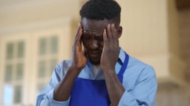 Aşırı yüklenmiş Afro-Amerikan erkek aşçı tapınakları ovalayarak içeride baş ağrısı çekiyor. Mutfakta migreni olan önlüklü üzgün aşçının yakın plan portresi..