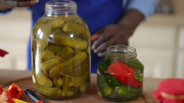 Frente vista garrafas com pepino orgânico e irreconhecível homem afro-americano fechando alimentos enlatados marinados com tampa. Jovem cara confiante conservando deliciosa refeição dentro de casa na cozinha. — Vídeo de Stock