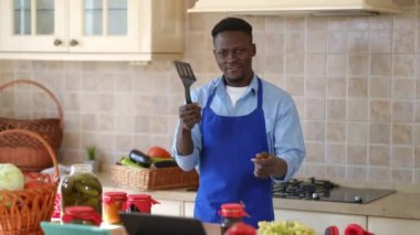 Önlüklü neşeli genç adam mutfakta dans ediyor ve sebzeleri terbiye ediyor. Neşeli, mutlu, Afro-Amerikan bir adamın portresi evde yemek konservesi yapıyor ve gülümsüyor..