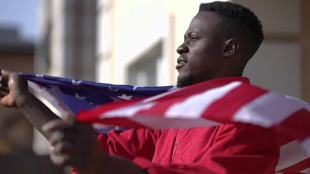 Side view proud confident African American man wrapping in USA flag standing in sunshine on backyard. Happy young guy enjoying sunny day outdoors in summer. — Stock Video
