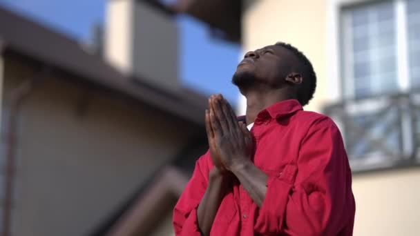 Joven afroamericano religioso con las manos apretadas rezando de pie a la luz del sol al aire libre. Esperando chico guapo agradeciendo a Dios disfrutando de un día de calma soleado en el patio trasero. — Vídeo de stock