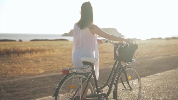 Panning lövés magabiztos nő sétál napsugárban kerékpárral a háttérben a Földközi-tenger és a mezőgazdasági területen Cipruson. Fiatal kaukázusi turista csodálja a természet séta szabadban naplementekor. — Stock videók