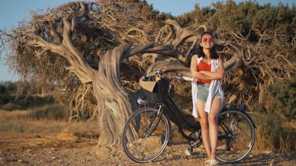 Bredt skudt portræt af fascineret glad ung kvinde stående med cykel ved træ på Cypern ved gylden solnedgang. Tilfredse smilende kaukasisk turist beundrer naturens skønhed nyder sommerferier. – Stock-video