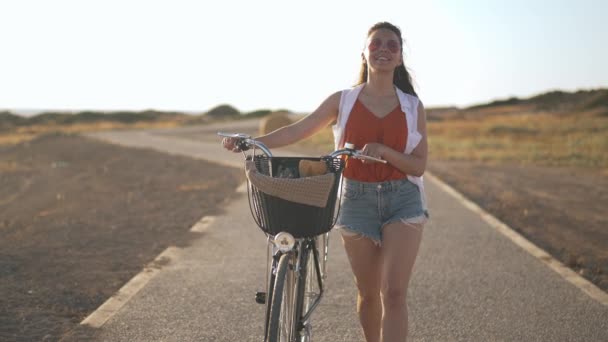 Vista frontal confiada delgada joven mujer paseando con bicicleta al atardecer en Chipre. Retrato de alegre hermosa sonriente turista caucásico disfrutando de vacaciones de verano mirando a su alrededor admirando la naturaleza. — Vídeo de stock