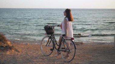 Bisikletle başka tarafa bakan kaygısız, mutlu genç bir kadın gülümseyerek Kıbrıs 'taki Akdeniz' in sarı sahilinde duruyor. Geniş açılı beyaz tenli turist gün batımında doğanın güzelliğine hayran..