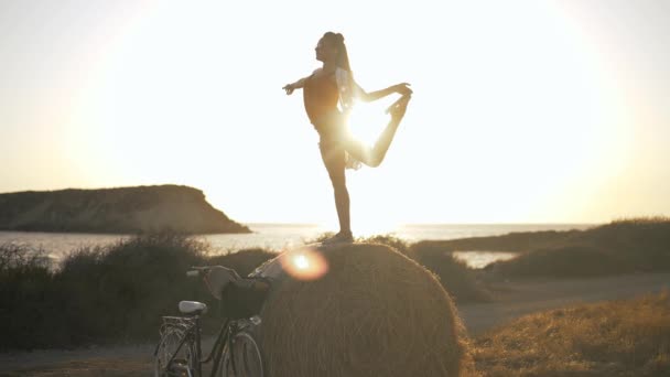 Широкий выстрел гибкий подходит кавказская молодая женщина поднимает ноги в солнечных лучах в замедленной съемке стоя на стоге сена на закате. Тонкая уверенная в себе дама, наслаждающаяся летним вечером на свежем воздухе на Cyprus. — стоковое видео