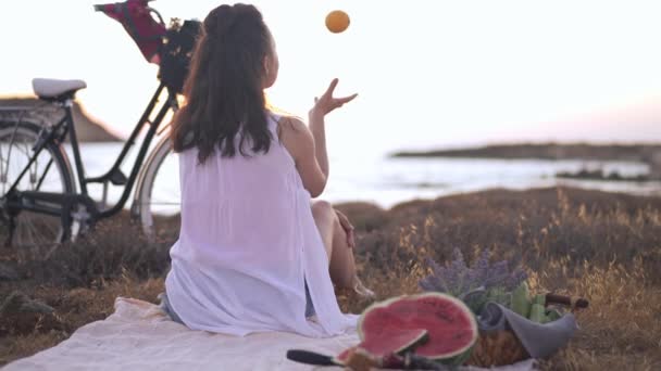 Widok z tyłu zrelaksowana szczupła kobieta żonglująca pomarańczą w zwolnionym tempie zwracając się do uśmiechniętej kamery. Szczęśliwy beztroski biały młody turysta pozowanie na piknik o zachodzie słońca na Cyprze. — Wideo stockowe