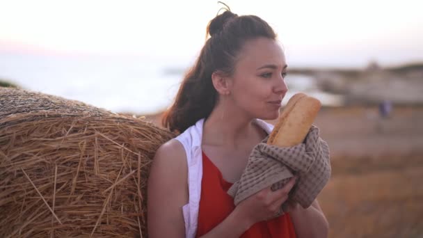 Szczupła piękna młoda kobieta pachnąca świeżo upieczonym bochenkiem chleba stojącym przy żółtym stogu siana na wiejskim polu. Portret pewnego siebie białego szczęśliwego rolnika na świeżym powietrzu na Cyprze o zachodzie słońca. — Wideo stockowe