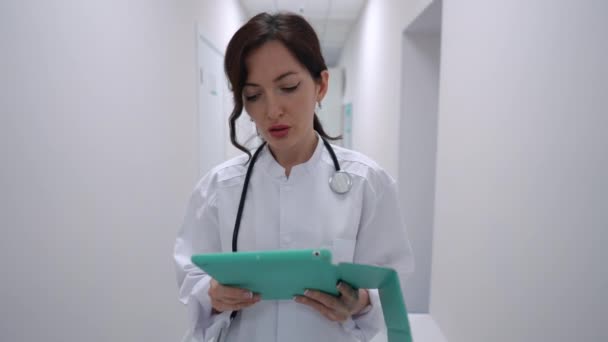 忧心忡忡的年轻女医生带着石碑慢吞吞地离去的画像 白种人医生带着网上测试结果在医院走廊里散步 — 图库视频影像