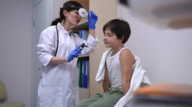 İnceleme koltuğunda oturan beyaz çocuk gülümseyen doktor olarak otoskopla kulakları kontrol ediyor. Hastanede kapalı alanda muayene olan kendinden emin küçük bir hastanın portresi.