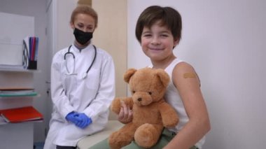 Coronavirus aşı merkezinde oyuncak ayıyla poz veren neşeli beyaz çocuk portresi. Omzunda yara bandıyla mutlu bir gülümseme ile hastanede bulanık bir doktorla oturan sevimli bir çocuk.