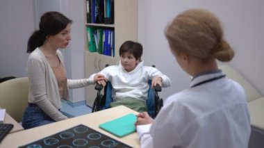 Tekerlekli sandalyedeki küçük beyaz çocuk endişeli bir kadınla hastanede doktorla konuşuyor. Düşünceli oğul ve endişeli anne klinikte danışmanlık uzmanı. Engellilik ve tıp konsepti