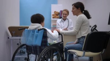 Arkaya bakınca gülümseyen beyaz bir anne tekerlekli sandalyede oğlunun elini tutarken arka planda oturan gülümseyen bir doktor gibi konuşuyor. Pozitif kadın, profesyonel danışmanlık uzmanı olan hasta çocuğu destekliyor.