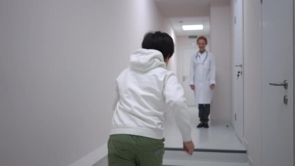 小児科医を抱きかかえる病院の廊下に立っている医者に走っている楽しい白人の少年の追跡ショット 病院で医者に会うことを楽しんでいる幸せな興奮した子供 スローモーション — ストック動画