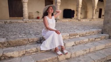 Geniş açılı mutlu beyaz elbiseli ve hasır şapkalı güzel kadın antik Kıbrıs şehrinde merdivenlerde oturmuş etrafına bakıyor. Yunan mimarisine hayran gülümseyen beyaz turist portresi