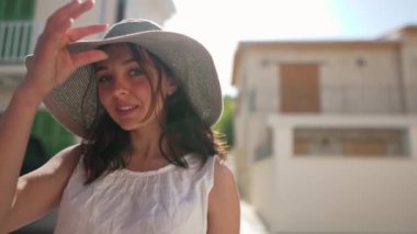 Günışığında gülümseyen genç bir kadının portresi Kıbrıs 'taki antik bir kasabanın arka planında kameraya bakıyor. Hasır şapkalı Kafkasyalı turist güneş ışınlarıyla dışarıda ağır çekimde poz veriyor.