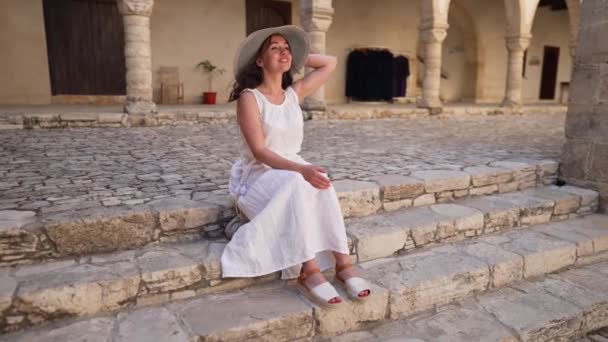白いドレスと藁帽子の広いショット幸せな美しい女性が周りを見て古代キプロスの街の階段に座っている 笑顔の肖像 ギリシャの建築を賞賛する白人観光客屋外 — ストック動画
