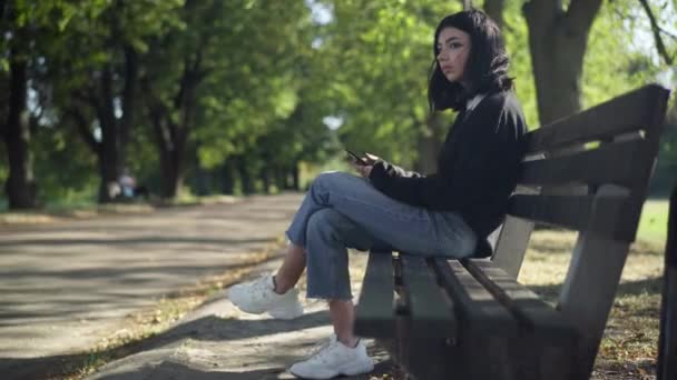 侧观白种人少女坐在长椅上的夏季公园冲浪社交媒体智能手机 图为周末在户外休息的自信青少年在网上发短信的全景 — 图库视频影像