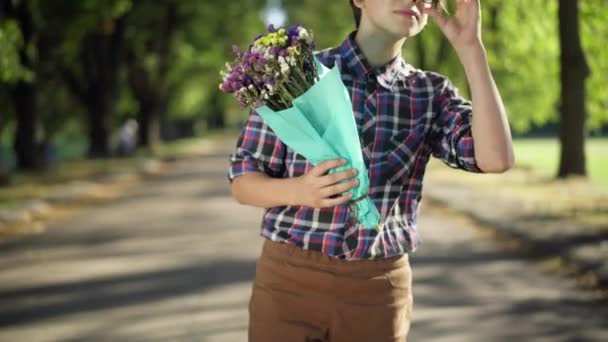 公園の路地に立っている花と恥ずかしがり屋のオタク10代の男の子はデートを待っています 認識できない知的白人のティーンエイジャーが花束を見て離れて見ている 最初の愛と不安 — ストック動画
