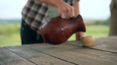 Tanımlanamayan bir adam, kadın eliyle bardağa kilden süt dökerek ön masaya süt şişesi koyuyor. Tarım arazilerinde sağlıklı süt içeceklerinin reklamı.