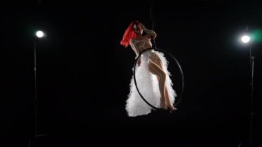 Esnek, baştan çıkarıcı, ince kadın arka ışıklandırmada hava çemberinin üzerinde duruyor beyaz eteği yavaş çekimde bükülerek atıyor. Siyah arka planda genç, beyaz saçlı, şehvetli jimnastikçinin görüntüsü.