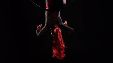 Yavaş çekimde dönen hava çemberinde baş aşağı asılı ince, esnek bir kadının yaya çekimi. Beyaz tenli gülümseyen jimnastikçi siyah arka planda sahnede poz verirken canlı kamera yükseliyor.