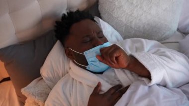 Hasta Afro-Amerikalı genç adamın Coronavirus maskesiyle yatakta öksürürken çekilmiş yakın plan portresi. Yatak odasında, Covid-19 belirtileri gösteren üzgün bir hastanın yüksek açılı görüntüsü. Salgın konsepti