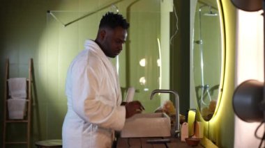 Yandan bakıldığında bornozlu yakışıklı Afro-Amerikan adam akıllı telefondan müzik açıp tıraş kremi sürüyor. Sabahları banyoda tıraş olan kendine güvenen bir gencin portresi.