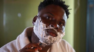 Yoğunlaştırılmış adamın tıraş olurken çekilen fotoğrafı. Tıraş bıçağıyla kameraya bakıyor. Odaklanmış Afrikalı Amerikalı gencin sabah rutinini evdeki banyoda yaparken yakın plan görüntüsü.