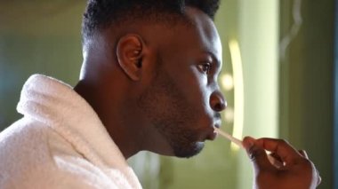 Evde tuvalette dişlerini fırçalayan konsantre bir adamın yan görünüşü. Odaklanmış Afro-Amerikalı genç adam sabahları ev işlerini yapıyor. Kişisel bakım ve diş sağlığı