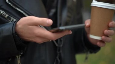 Elinde kağıt kahve fincanıyla sosyal medyada gezinen beyaz bir gencin yakın plan akıllı telefonu. Deri ceketli, kendine güveni olmayan bir adam internette mesajlaşarak sörf yapıyor.