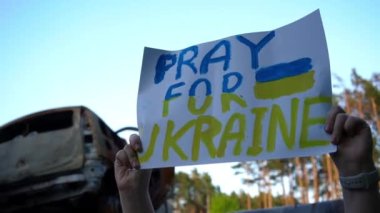 Beyaz kadın ellerinde sarı ve mavi bayrak renginde Ukrayna levhası için yakın plan dua edin. Yanmış bombalanmış arabaların önünde yardım isteyen tanınmayan Ukraynalı genç bir kadın.