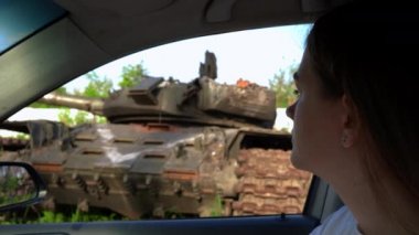 Yan manzaralı genç bir kadın arabada oturmuş Ukrayna 'da yanan tanklara bakıyor ve başka tarafa bakıyor. Sağdaki Ukraynalı kadın arka planda düşman askeri araçlarıyla yok edildi.