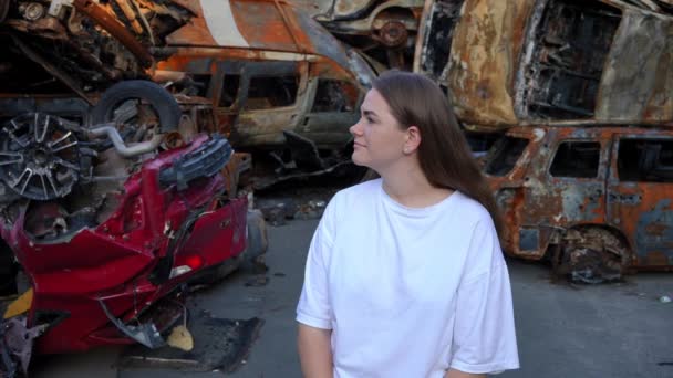 在战区的汽车垃圾堆上 看着周围被毁的汽车 脸上带着悲伤的表情 把目光投向年轻 体贴的女人 美丽的黑发乌克兰女人站在破车前思考 — 图库视频影像