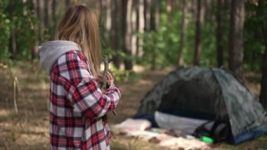 Kamp çadırına bakan güzel bir kadın elinde odun dallarıyla kameraya bakarak el sallıyor. Keyifli bir turistin portresi kamp yapıyor.