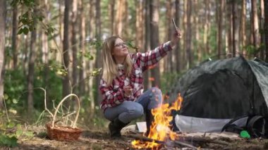Canlı kamera, şenlik ateşinde video blogu çekerken mantar kızartan büyüleyici güzel kadın olarak soldan sağa hareket ediyor. Genç kendine güvenen beyaz blogcu ormanda kamp yaparken gülümsüyor