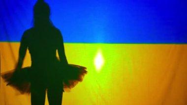 Silhouette of ballerina spinning dancing at background of Ukrainian flag with backlit. Slim unrecognizable woman in tutu performing dance. Art and war concept