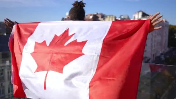 回头看 拿着加拿大国旗的年轻人在户外阳光下欣赏城市 自信快乐的非裔美国人 国旗高悬在阳光下 — 图库视频影像
