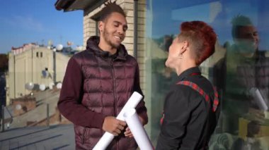 Afro-Amerikalı bir adam ve beyaz bir kadının terastaki cam çitlere doğru ağır çekimde yürürken görüntüsü. Güler yüzlü müteahhit ve müşteri şehir merkezinde gün ışığında dolaşıyorlar.