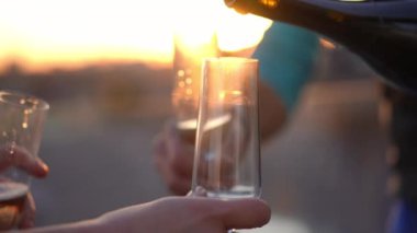 Close-up pouring champagne in glasses in sunrays at golden sunset. Unrecognizable group of Caucasian people celebrating enjoying leisure on roof in urban city at sundown