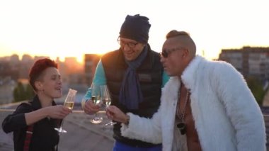 Group of cheerful Caucasian friends toasting drinking champagne on roof in urban city at sunset. Happy joyful men and woman clinking glasses enjoying refreshing beverage outdoors