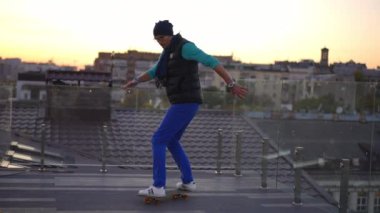 Wide shot adult man training skateboarding on roof in urban city riding to camera leaving. Concentrated Caucasian guy at sunset on rooftop enjoying hobby. Lifestyle and sport concept