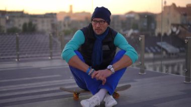 Positive man sitting on skateboard singing dancing with urban city at sunset at background. Portrait of carefree Caucasian guy enjoying evening outdoors