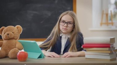 Dissatisfied Caucasian schoolgirl gesturing thumbs down looking at camera with tired facial expression. Medium shot portrait of exhausted overburdened student posing in classroom in school indoors
