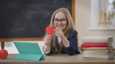 Happy satisfied teenage schoolgirl posing with heart shape card in classroom. Portrait of cheerful positive Caucasian girl looking at camera smiling sitting at desk with tablet and books