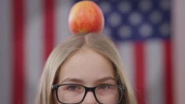 Gözlüklü, kafasında elma, arka planında Amerikan bayrağı olan liseli kızların yakın çekimleri. Neşeli kaygısız mutlu kız kameraya bakıyor. Eğitim ve sağlıklı beslenme kavramı