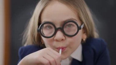 Headshot of nerd schoolgirl in high dioptres eyeglasses drinking juice with straw looking at camera. front view close-up of confident intelligent Caucasian student posing in school classroom